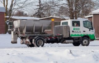 Small truck with a snow spreader parked in a snowy area near buildings and trees.